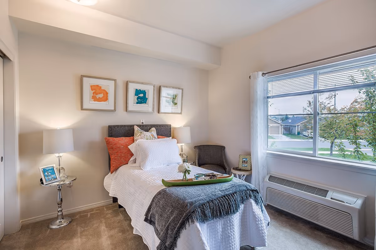 A cozy bedroom with a single bed dressed in white bedding and a gray throw blanket. The bed has multiple pillows including a red one and a floral one. On the wall above the bed are three framed abstract art pieces in orange, blue, and green. There are two bedside tables with lamps, a small chair, and a window with white curtains showing a view of houses and trees outside.