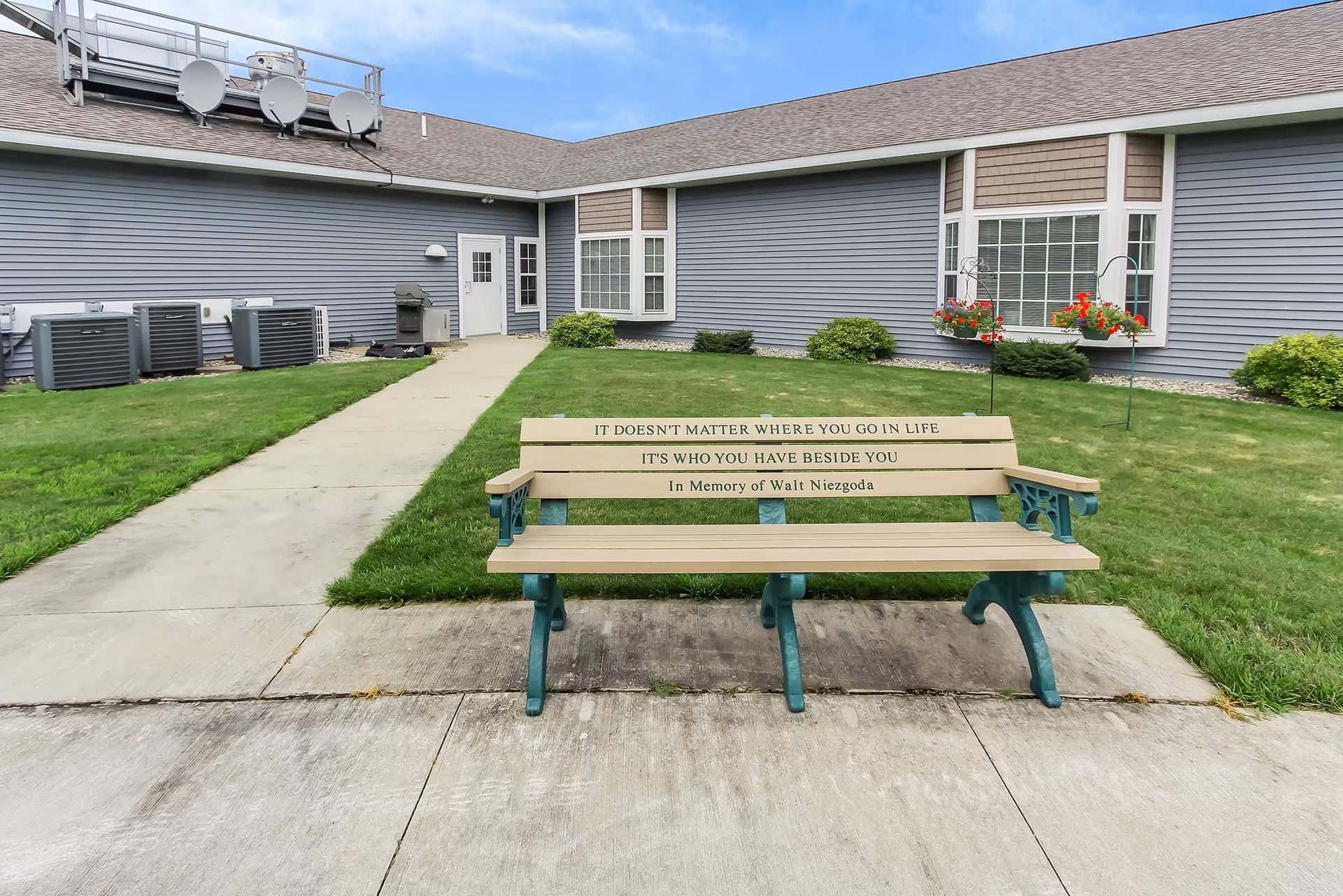 Outdoor view of a courtyard area at White Lake Assisted Living and Memory Care featuring a memorial bench with the inscription 'It doesn't matter where you go in life, it's who you have beside you. In memory of Walt Niezgoda.' The bench is placed on a concrete slab with a sidewalk leading to a building with gray siding, windows, and a door. There are green lawns, bushes, and hanging flower baskets near the building.