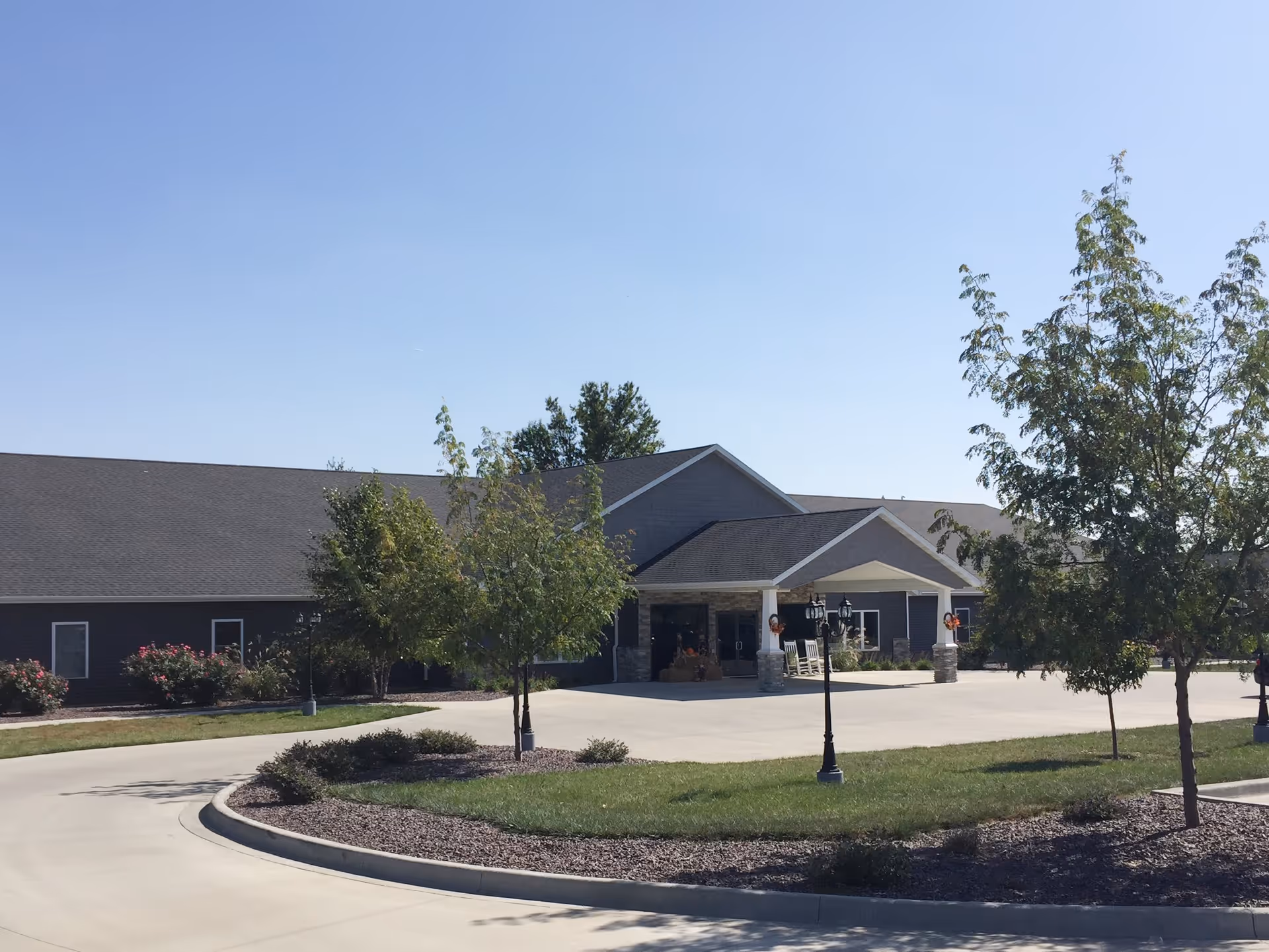 Exterior view of a single-story senior living facility building with a covered entrance, surrounded by a paved driveway, green grass, trees, and landscaping under a clear blue sky.