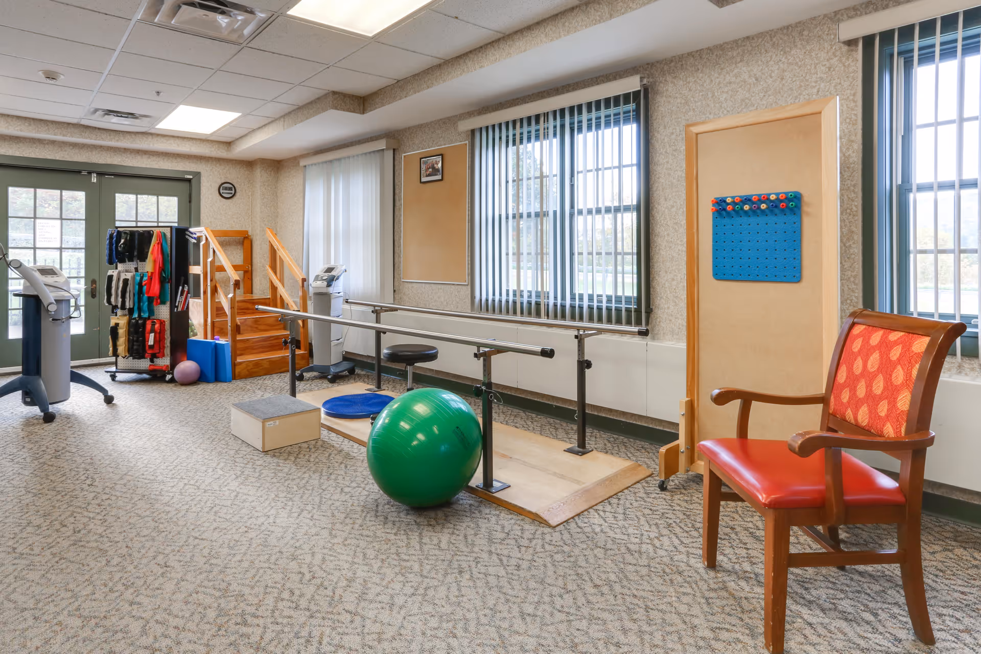 A physical therapy or rehabilitation room with exercise equipment including parallel bars, a green exercise ball, balance pads, a small wooden step platform, and a rack with various exercise bands and weights. There is a wooden chair with red upholstery and a pegboard on a stand near the windows with vertical blinds. The room has carpeted flooring and fluorescent ceiling lights.