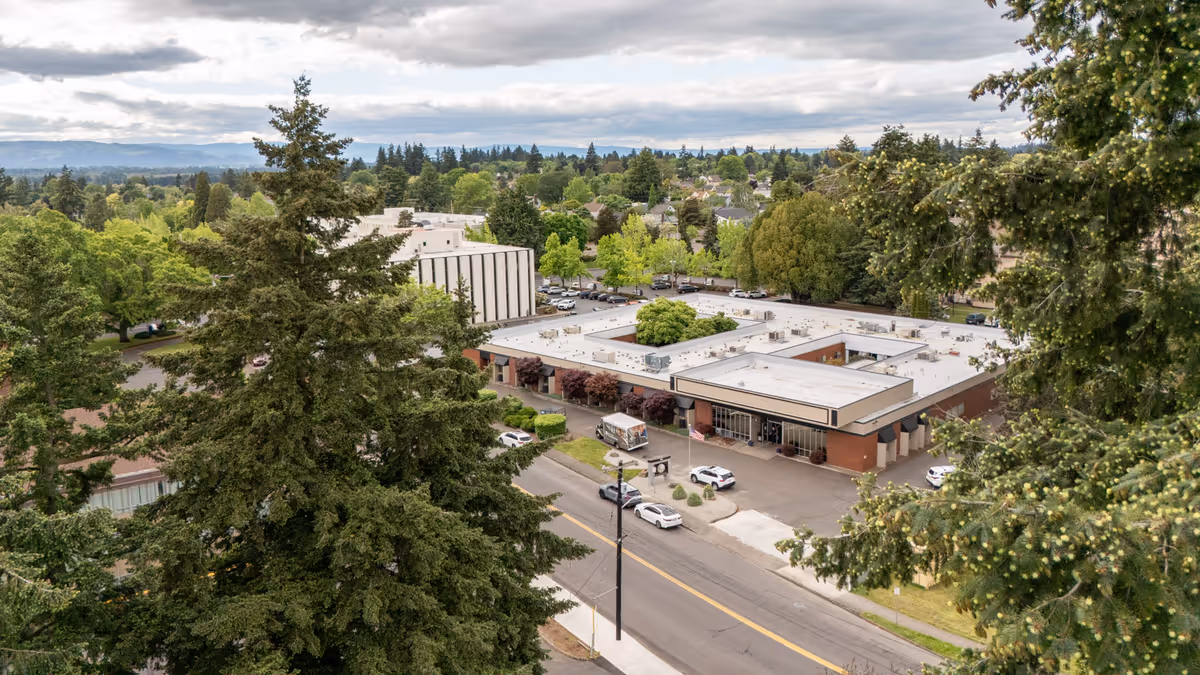 Aerial view of a single-story brick building with a parking lot and tree-lined surroundings in a residential neighborhood.