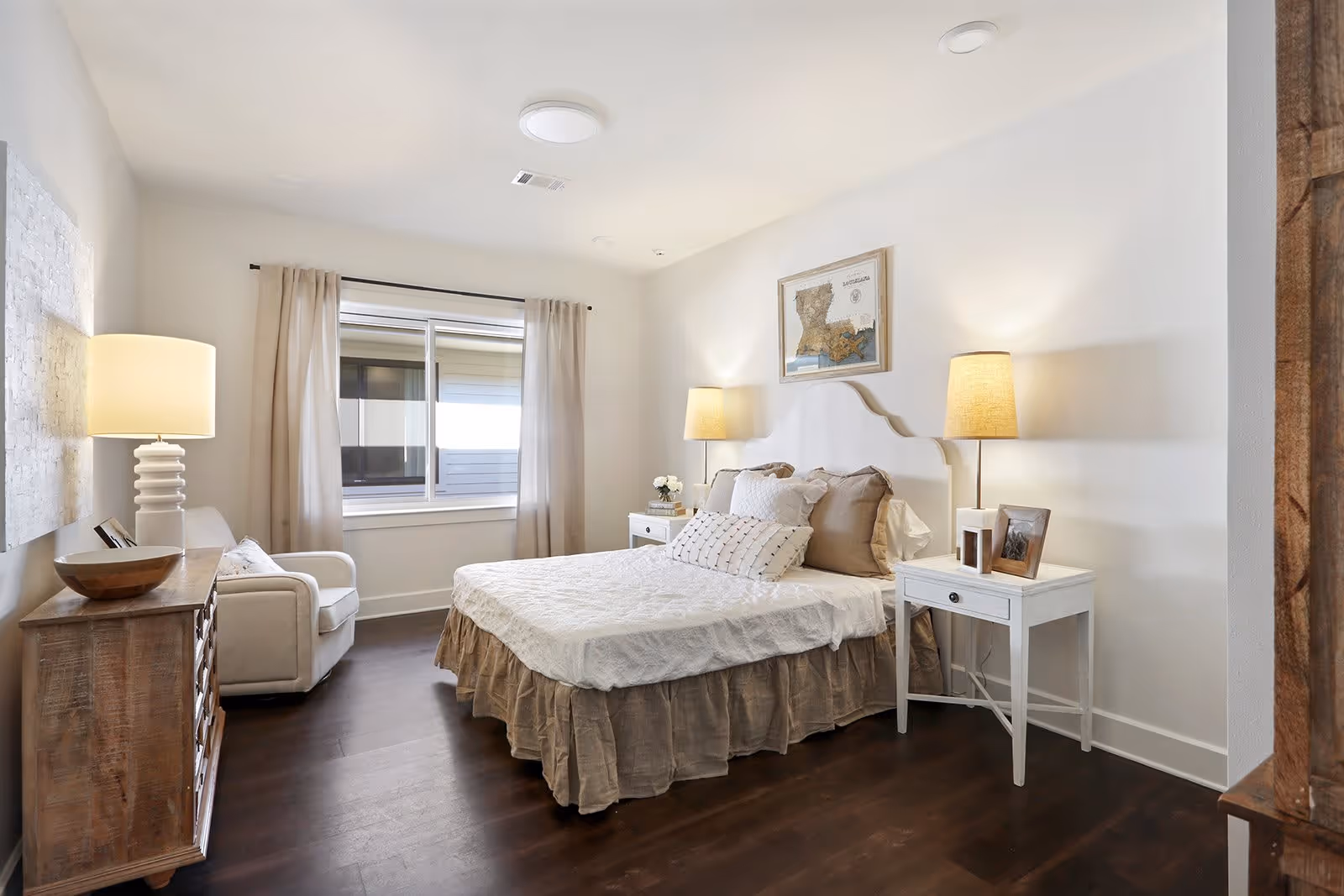 A bright and cozy bedroom with a large bed dressed in white and beige linens, two bedside tables each with a lamp, a wooden dresser with a lamp and decorative bowl, a comfortable armchair near a window with beige curtains, and a framed map artwork on the wall above the bed.