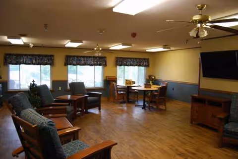 Bright senior living common room with seating, small tables, a wall TV, and windows.