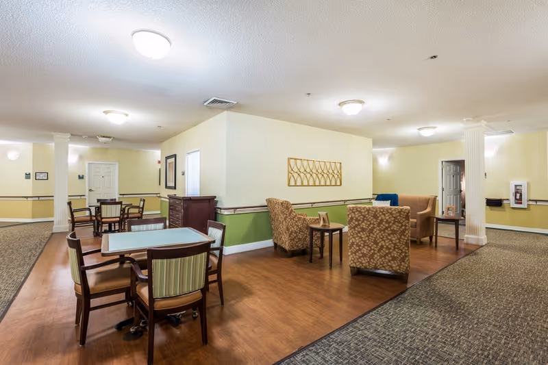 Communal sitting and dining area in a senior living facility with tables, chairs, and upholstered armchairs.