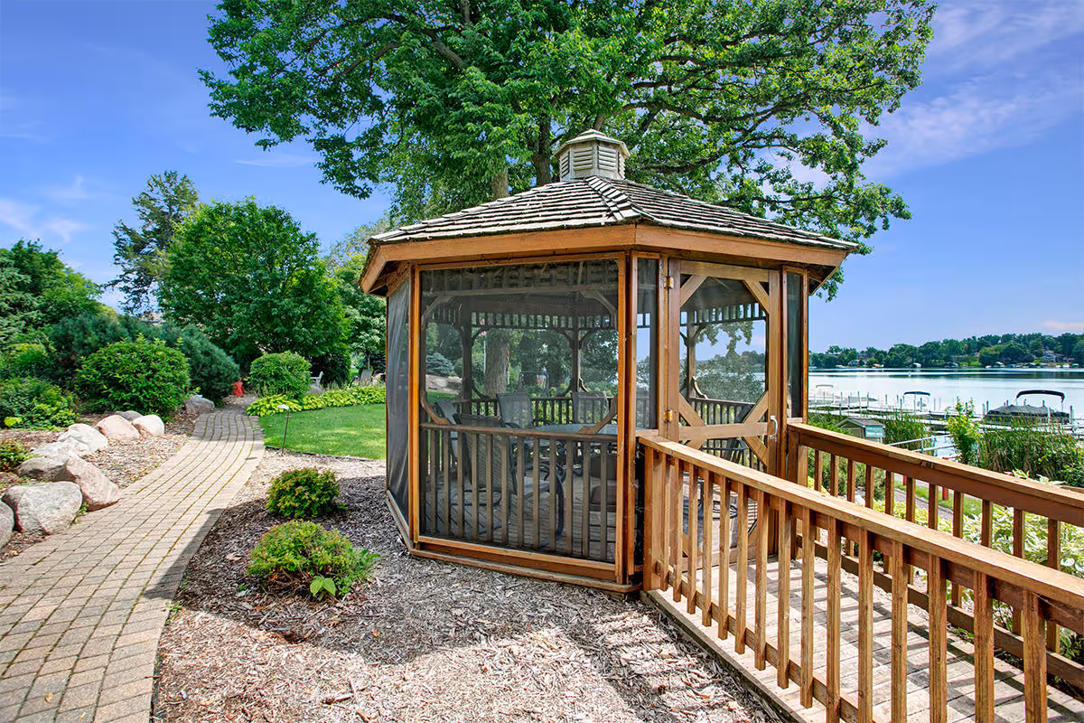 A wooden gazebo with screened windows and a shingled roof sits next to a paved walkway surrounded by landscaped greenery and rocks. In the background, there is a lake with several boats docked and trees under a clear blue sky.