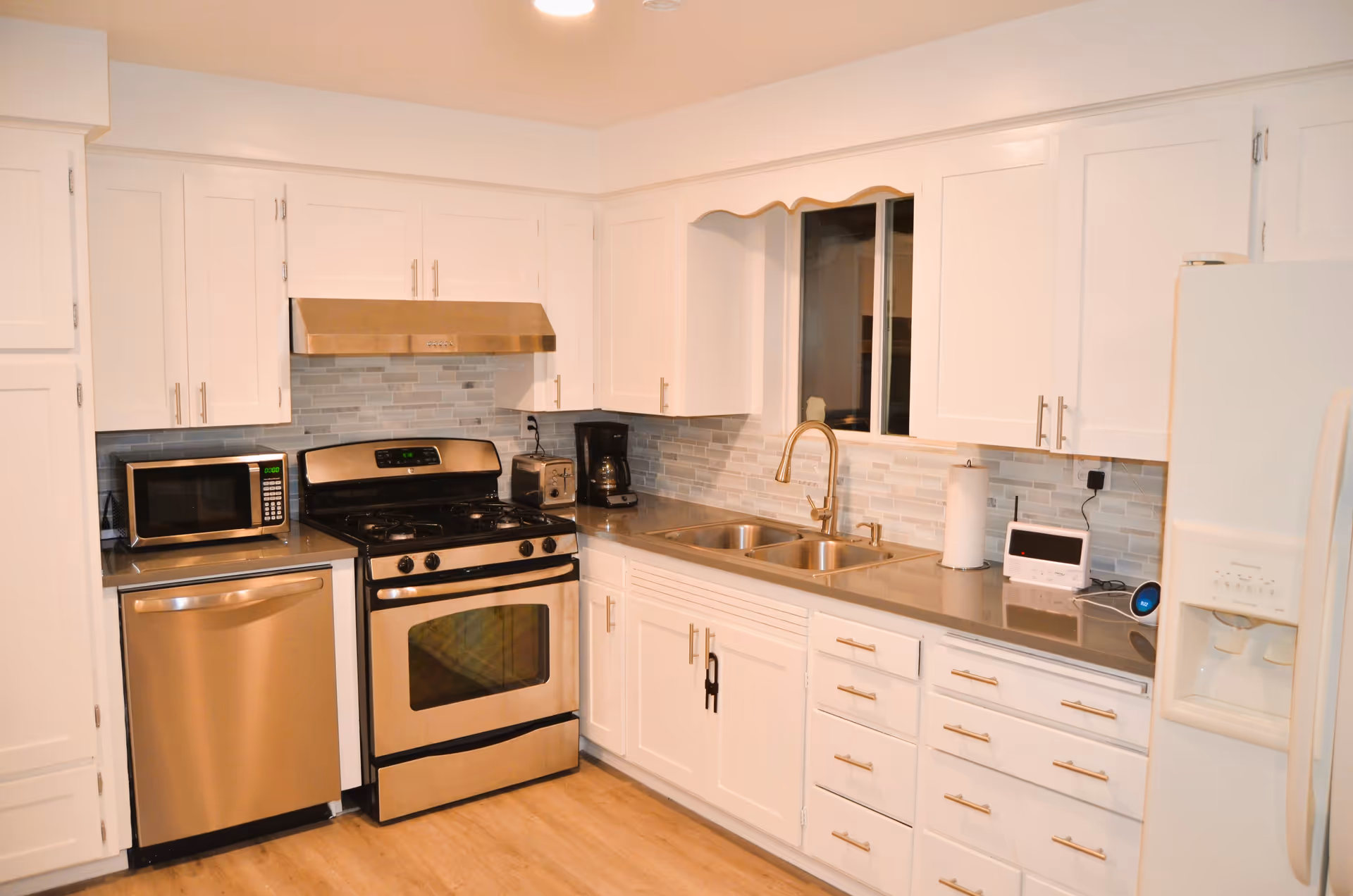 A modern kitchen with white cabinets and drawers, stainless steel appliances including a dishwasher, stove, microwave, and refrigerator. There is a double sink with a gold-colored faucet, a coffee maker, toaster, paper towel holder, and a small electronic device on the countertop. The backsplash is made of light gray and white tiles, and the floor is wooden.