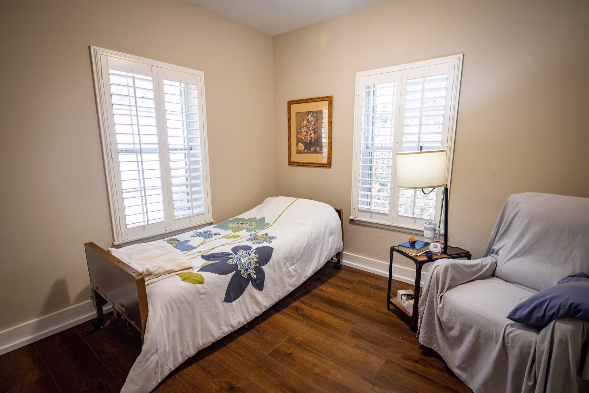 A simple bedroom with a single bed, armchair, side table with a lamp, and two shuttered windows.