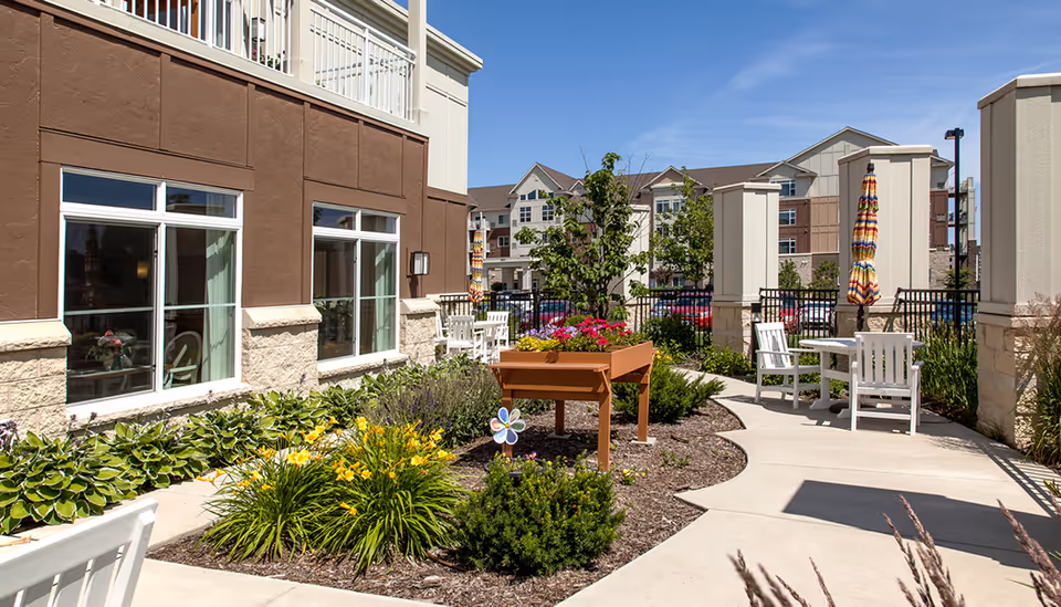Outdoor patio area at The Waters on Mayowood featuring a paved walkway, garden beds with flowers and shrubs, white chairs and tables with colorful umbrellas, and a multi-story building in the background under a clear blue sky.
