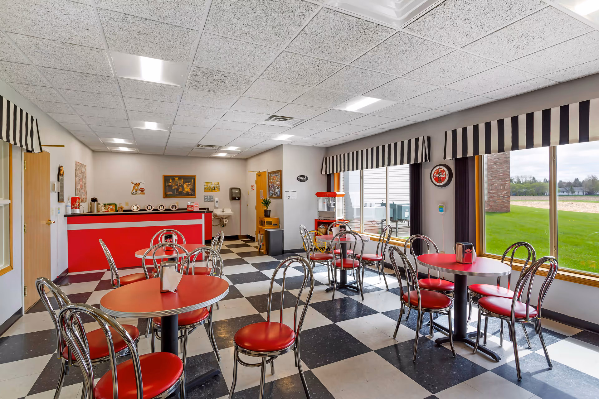 Bright retro-style dining room with red round tables, chrome chairs, a checkered floor and a service counter near large windows.