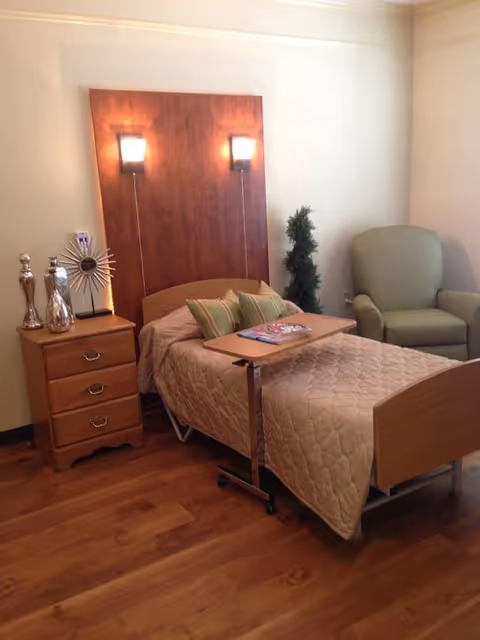 A cozy bedroom in a senior living facility featuring a single bed with a wooden headboard and beige quilted bedspread. The bed has a small overbed table with magazines on it. Next to the bed is a wooden nightstand with decorative items and a modern clock. There are two wall-mounted lights above the headboard. A green upholstered armchair and a small artificial plant are placed in the corner of the room. The floor is wooden, and the walls are painted light beige.