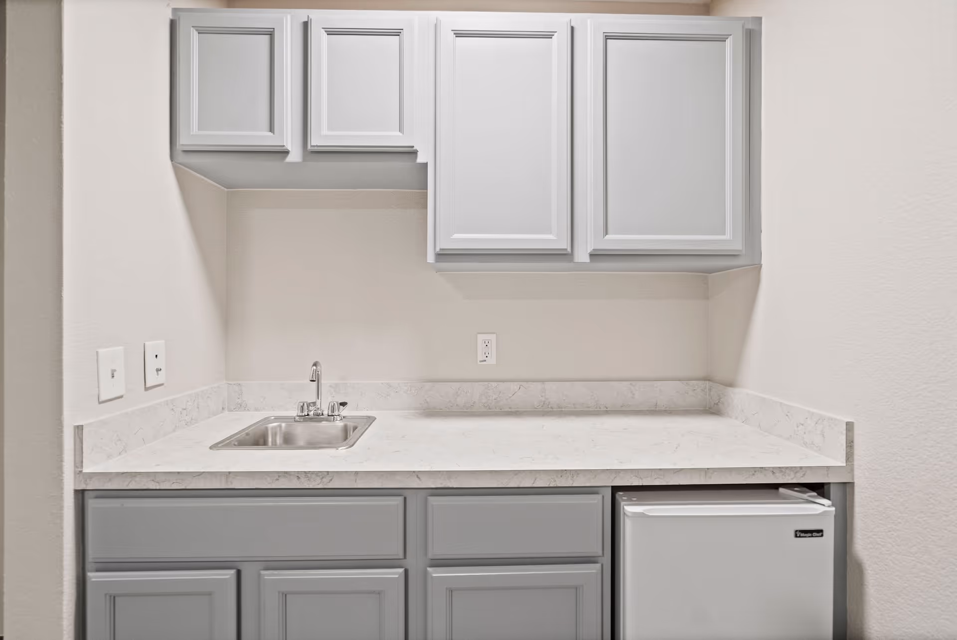 A small kitchenette area with light gray upper and lower cabinets, a white marble countertop, a small stainless steel sink with a faucet, and a compact white refrigerator underneath the counter on the right side.