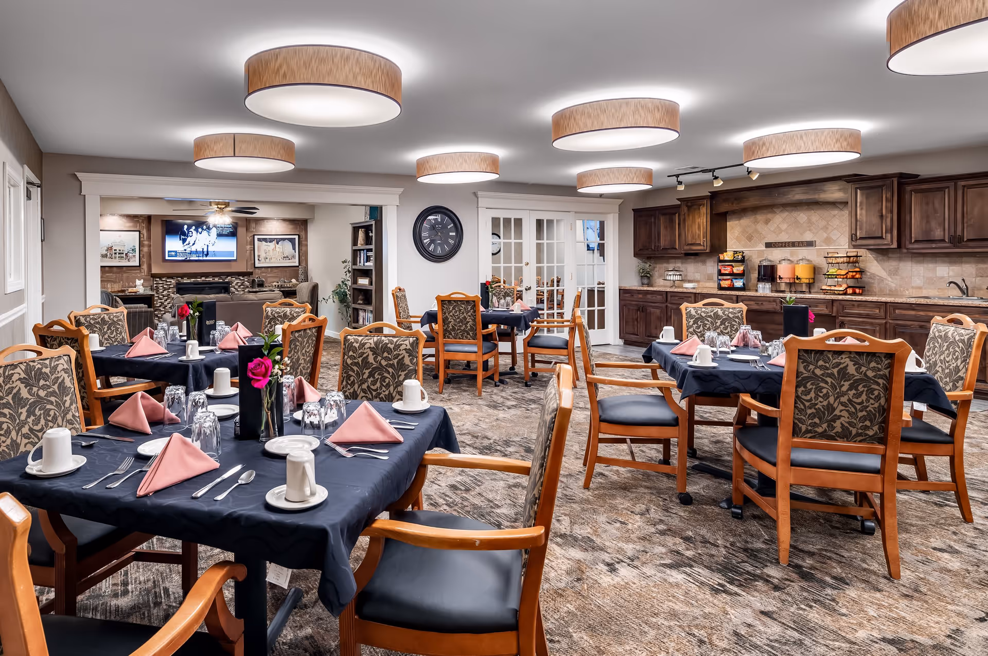 A dining room in a senior living facility with several tables covered in dark tablecloths, each set with pink folded napkins, white cups, glasses, and silverware. The chairs have wooden frames with patterned upholstery. The room features modern circular ceiling lights, a carpeted floor, and a kitchenette area with wooden cabinets and a beverage station. In the background, there is a cozy sitting area with a TV mounted above a fireplace.
