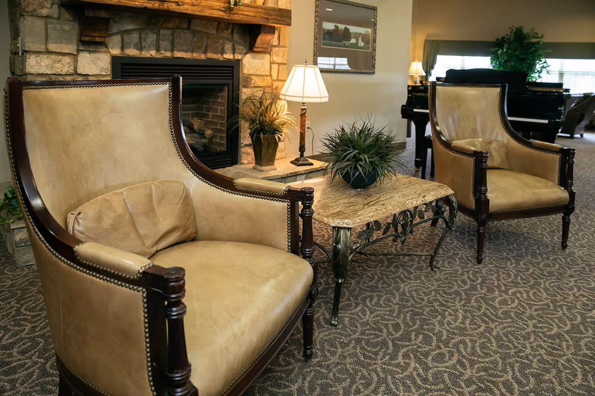 A cozy living room area with two beige leather armchairs with wooden frames, a small ornate coffee table with a potted plant, a stone fireplace, a table lamp, and a piano in the background near a window with blinds.
