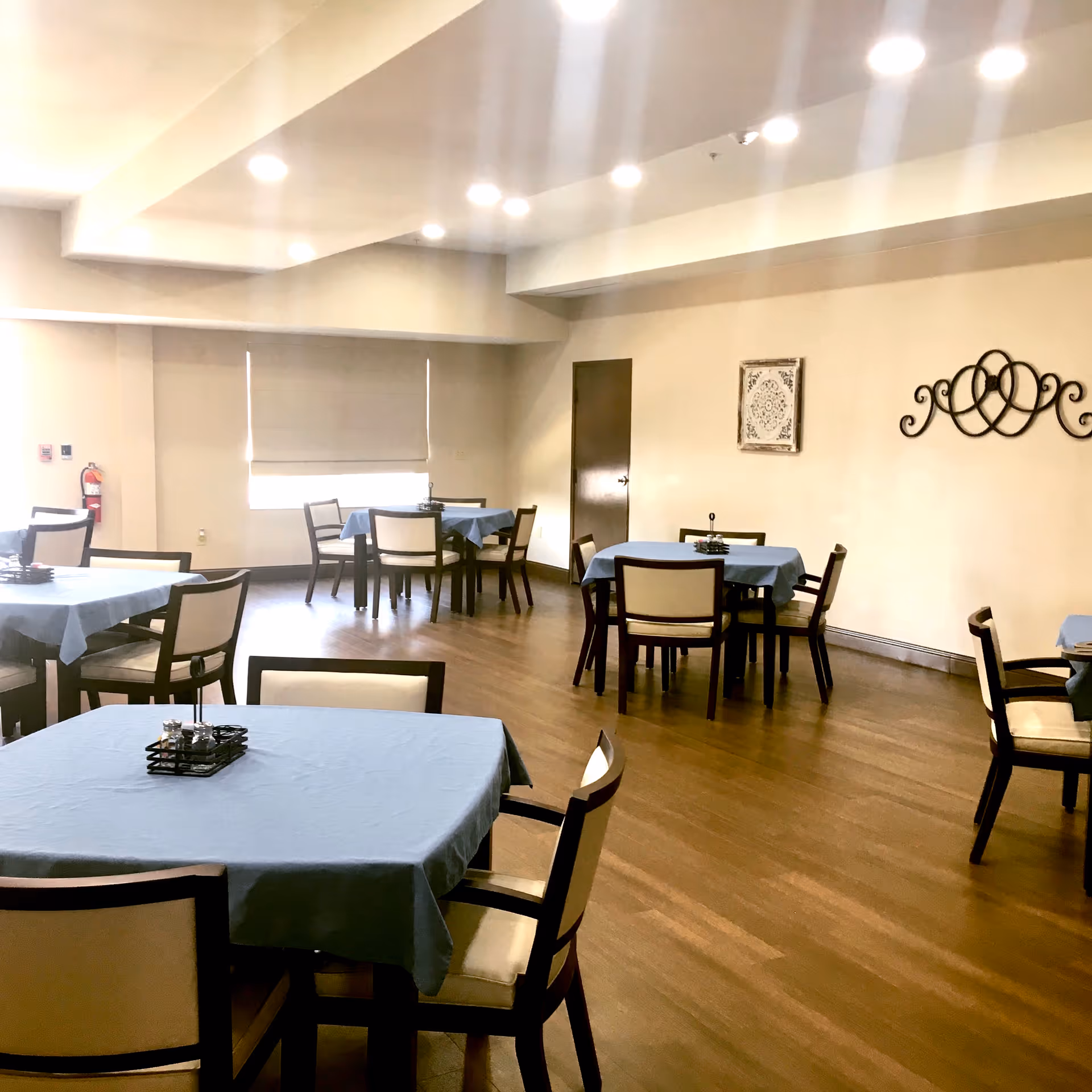 A dining room with several square tables covered with light blue tablecloths, each surrounded by four chairs with cream-colored cushions. The room has wooden flooring, beige walls, ceiling lights, a window with a closed blind, a fire extinguisher on the left wall, and decorative wall art on the right wall.