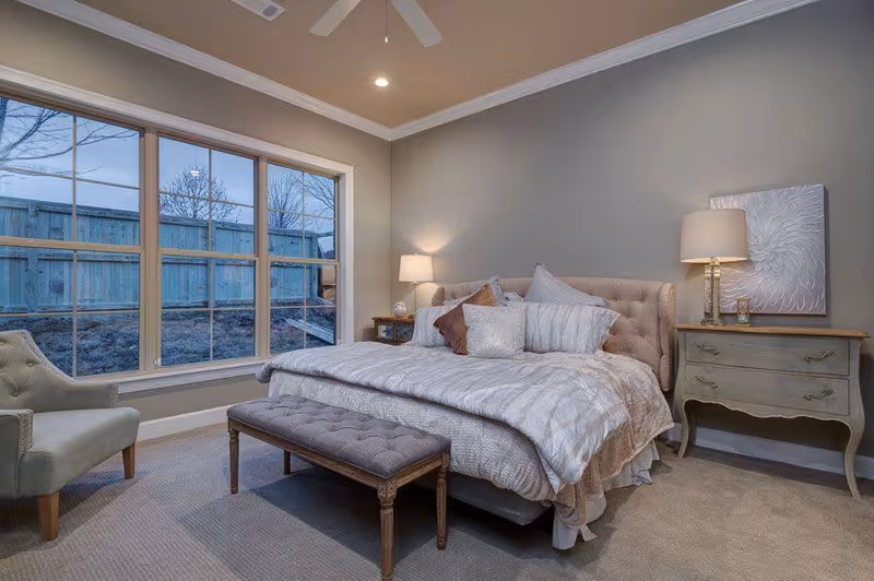 A cozy bedroom with a large bed featuring multiple pillows and a tufted headboard. There is a cushioned bench at the foot of the bed, a light green armchair near a large window with a view of a fenced yard, and two bedside tables each with a lamp. The walls are painted gray and the room has beige carpeting.