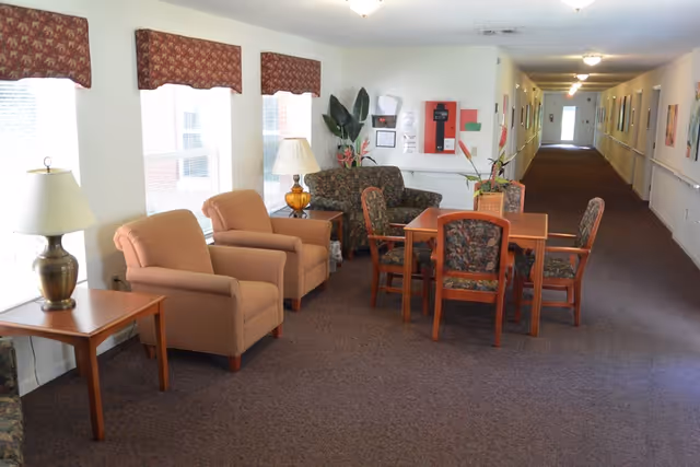 A well-lit hallway in an assisted living facility with several windows on the left side covered by patterned valances. The hallway features a seating area with two beige armchairs, a wooden side table with a lamp, a wooden table with four floral-patterned chairs, and a floral-patterned loveseat. There are plants and a fire extinguisher mounted on the wall in the background, and the hallway extends into the distance with handrails along the walls and framed pictures hanging.
