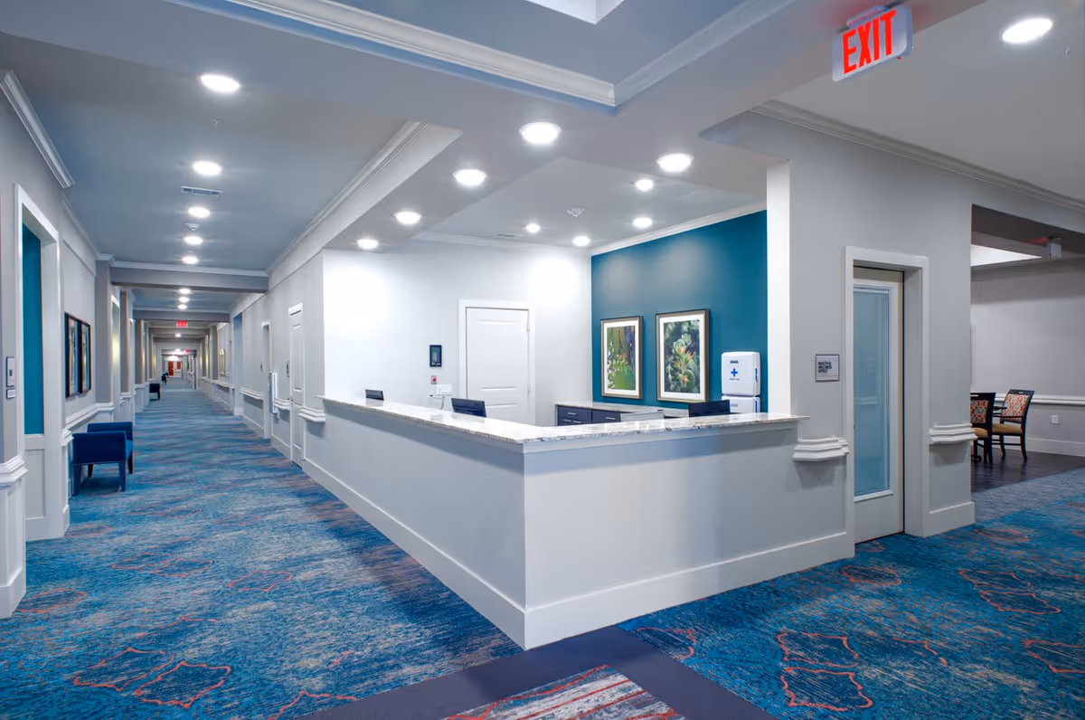 Interior view of a senior living facility hallway with a reception desk on the right side. The hallway has blue patterned carpet, white walls, and ceiling lights. There are framed pictures on the walls and an exit sign above a doorway. Chairs are placed along the hallway and a small seating area with chairs and a table is visible in the background.