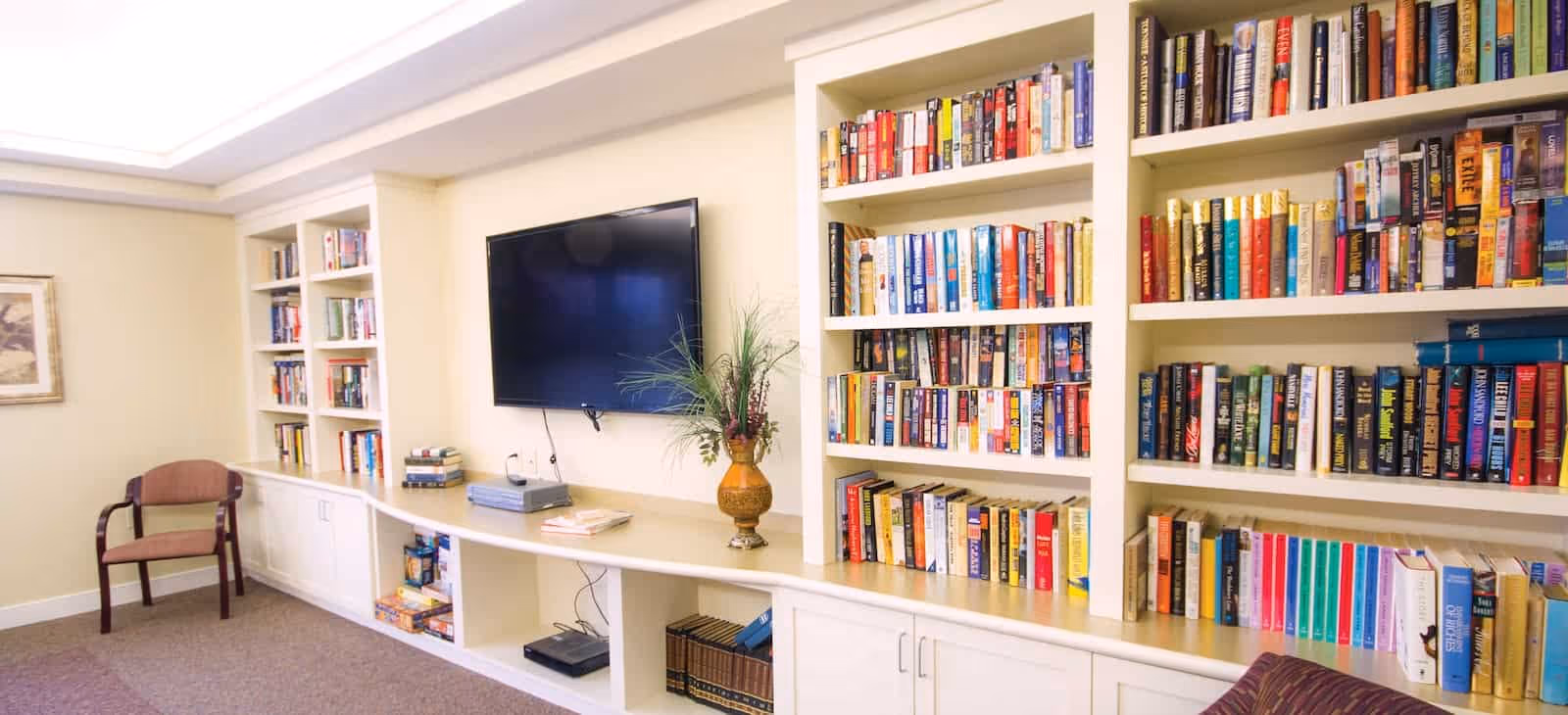 A cozy interior room with built-in white bookshelves filled with books, a flat-screen TV mounted on the wall, a decorative vase with plants on the countertop, and a wooden chair with armrests placed against the wall.
