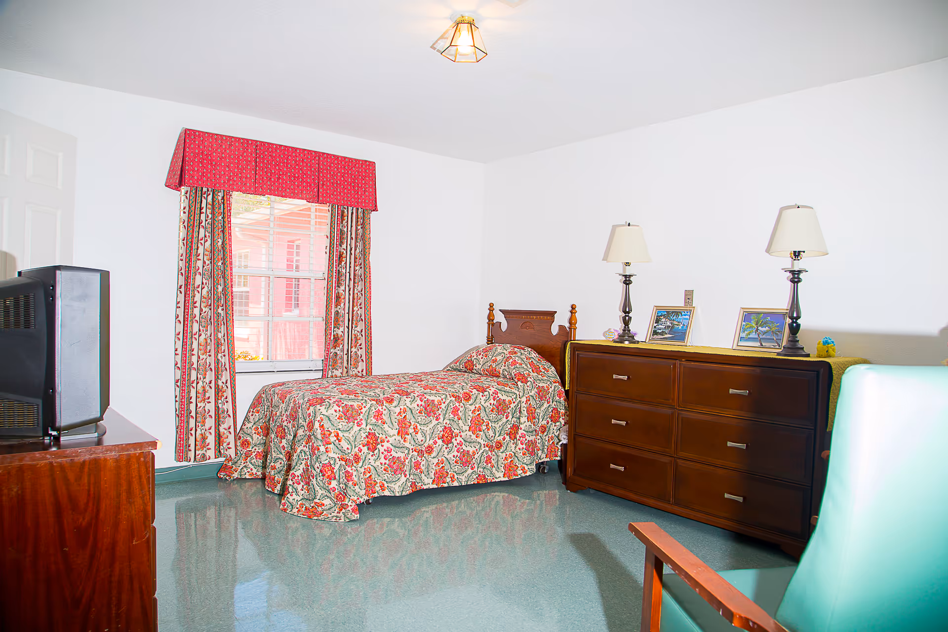 A small bedroom with a single bed covered in a floral bedspread, a wooden dresser with two lamps and framed pictures on top, a green armchair, and a window with floral curtains and a red valance. There is also an older style television on a wooden stand.