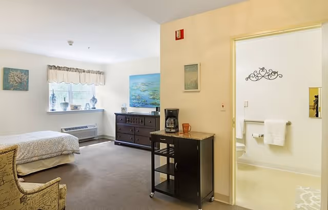 A bright senior living facility room with a bed on the left, a window with decorative vases and a valance, a dark wooden dresser with a painting above it, and a small black cart with a coffee maker and mug. To the right, there is an open doorway leading to a bathroom with white walls, a towel rack with white towels, and a decorative wall piece above the toilet.