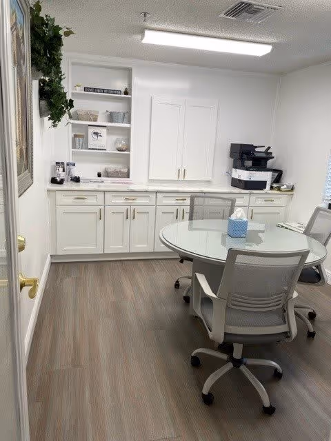 Small interior meeting/office room with a round glass table and rolling chairs, white cabinets and shelving with a printer and decor.