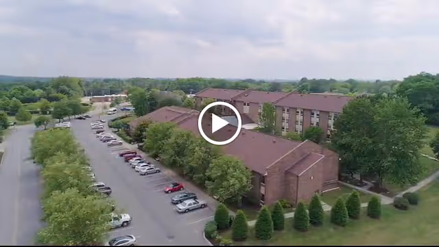 Aerial view of Park Place Retirement Community showing multiple brick buildings with brown roofs surrounded by green trees and landscaped bushes. There is a parking lot with several cars parked along the side of the buildings. The sky is partly cloudy.