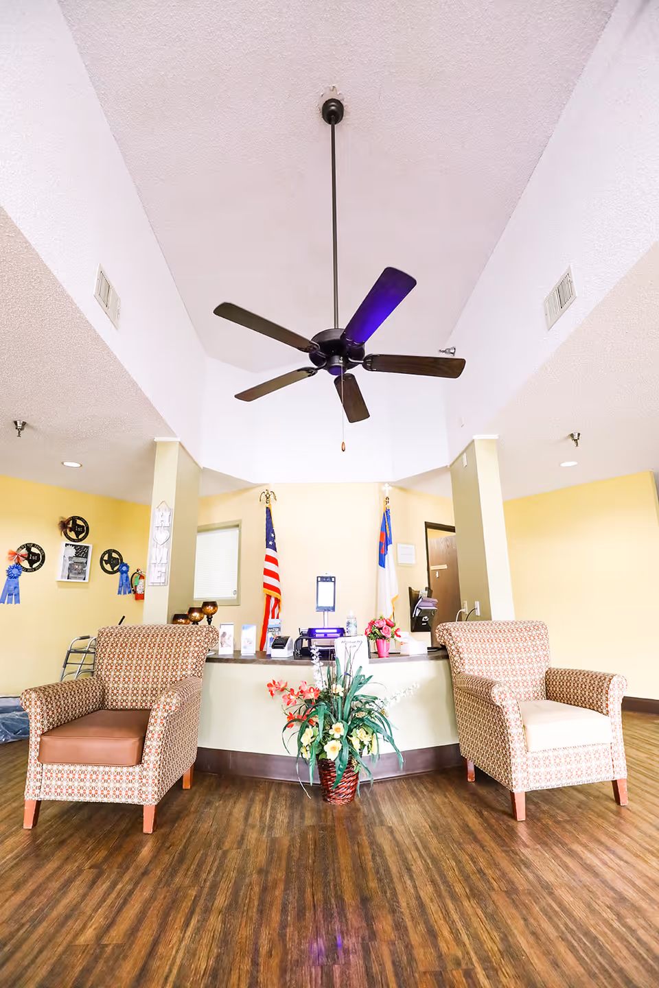 Reception area with two patterned armchairs, a central desk with flags and a flower arrangement, and a ceiling fan overhead.