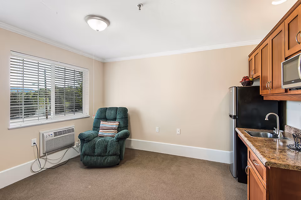 A small living area with a green recliner chair featuring a striped pillow, next to a window with white blinds and an air conditioning unit below it. To the right, there is a kitchenette with wooden cabinets, a granite countertop, a stainless steel sink, a black refrigerator, and a microwave. The walls are beige and the floor is carpeted.