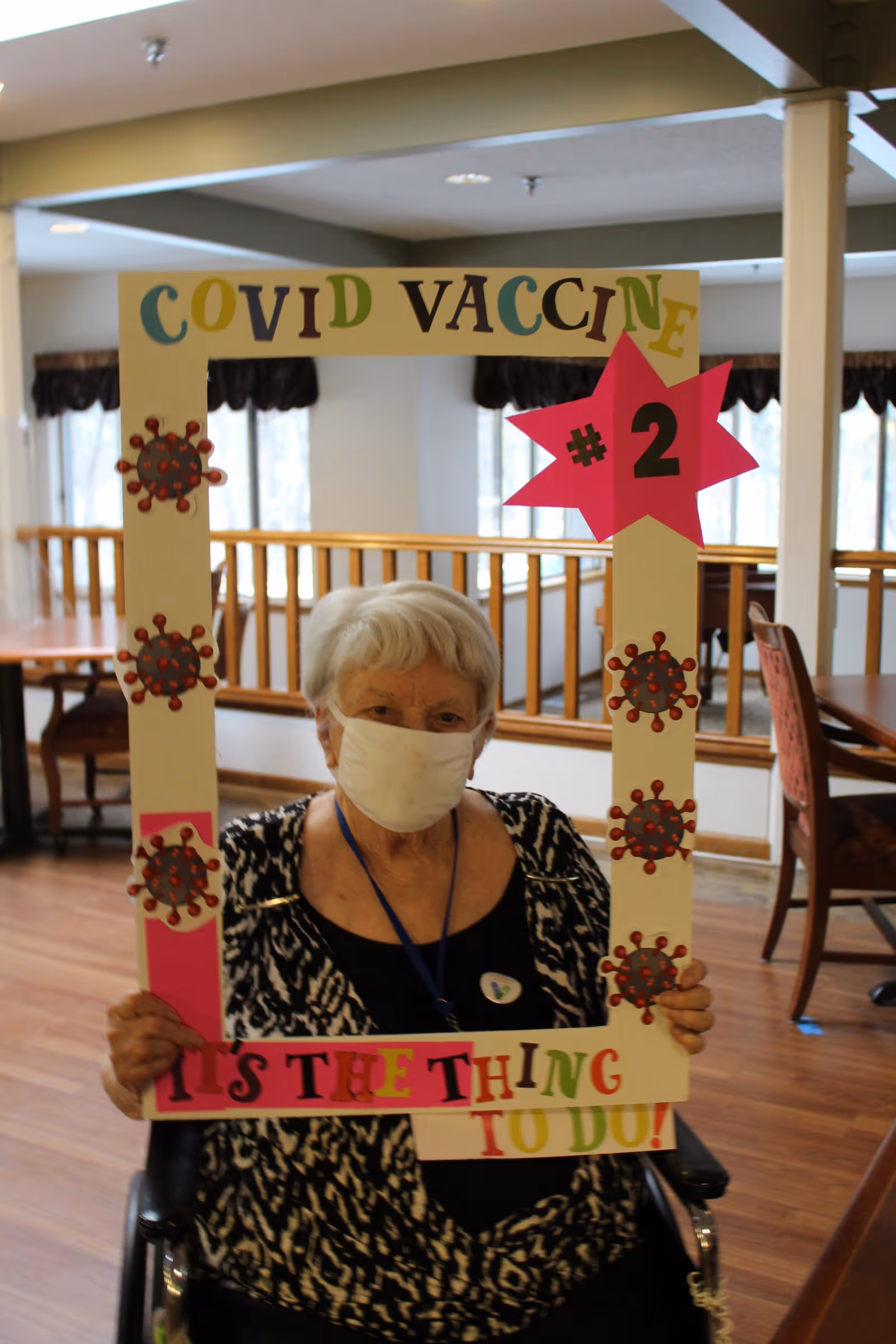 An elderly woman wearing a face mask sits in a wheelchair inside a room with wooden floors and tables. She is holding a large decorated frame that reads 'COVID VACCINE #2' and 'IT'S THE THING TO DO!' with images of virus particles on the frame.