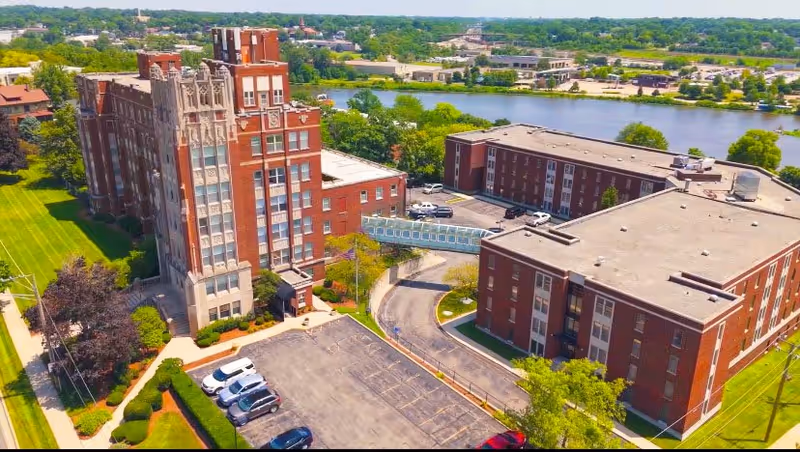 Aerial view of Living Fox Knoll Village showing multiple red brick buildings connected by a skywalk, surrounded by parking lots, green lawns, trees, and a river in the background under a clear sky.