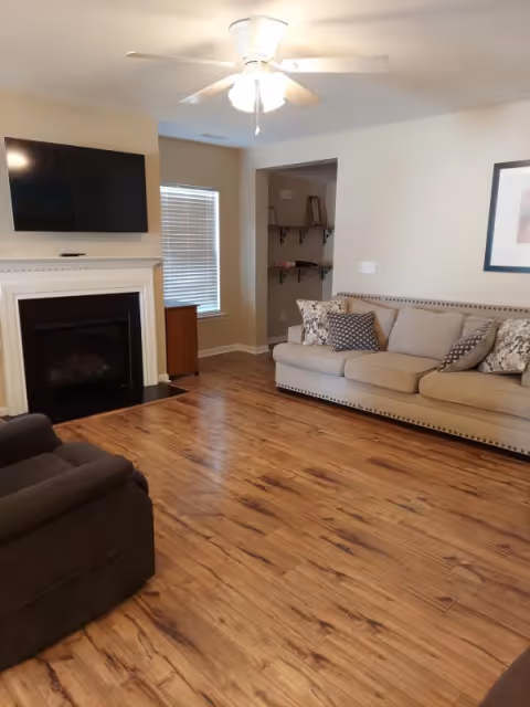 A cozy living room with wooden flooring, a beige sofa with patterned cushions, a dark armchair, a white ceiling fan with light, a wall-mounted flat-screen TV above a white fireplace, and a window with blinds.
