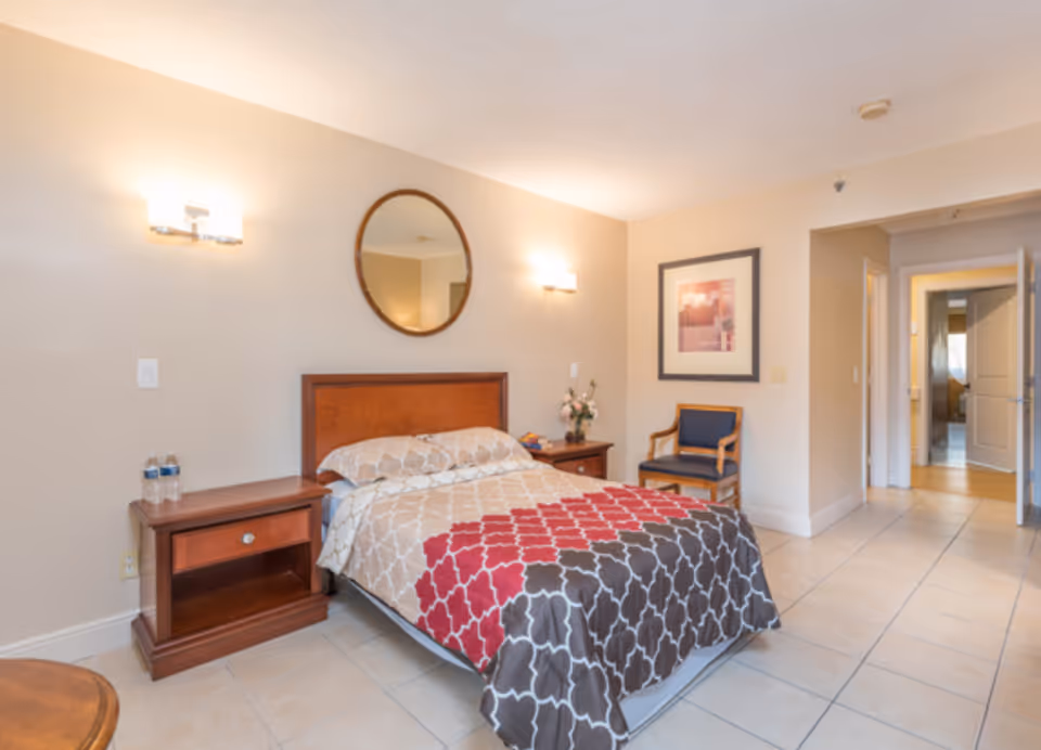 A bright assisted-living bedroom with a bed dressed in a patterned comforter, wooden nightstands, a round wall mirror, a chair and an open doorway.