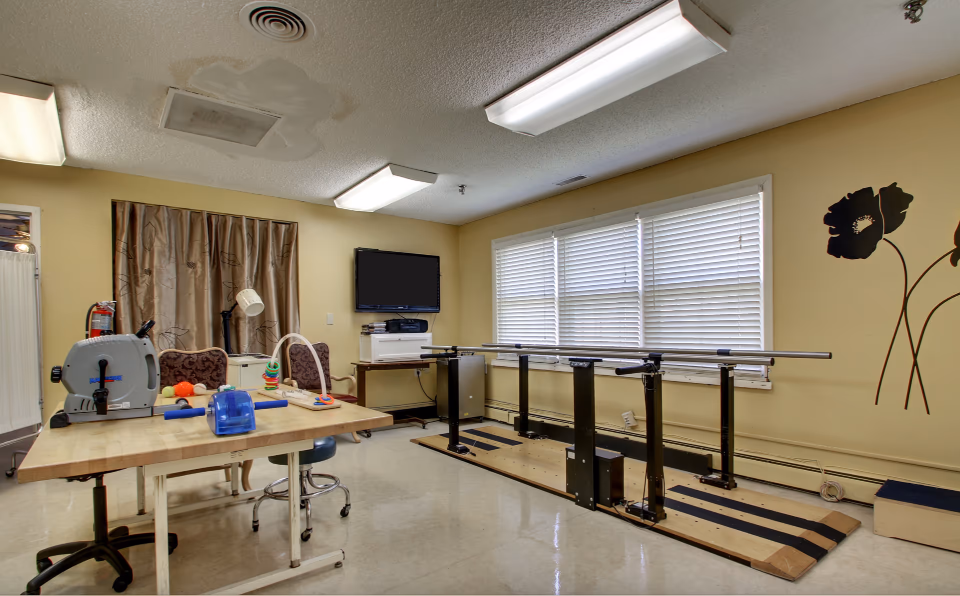 A therapy or rehabilitation room with exercise equipment including a stationary bike, parallel bars for walking practice, and a table with therapy tools. The room has beige walls, a window with blinds, a wall-mounted TV, and floral wall decals.