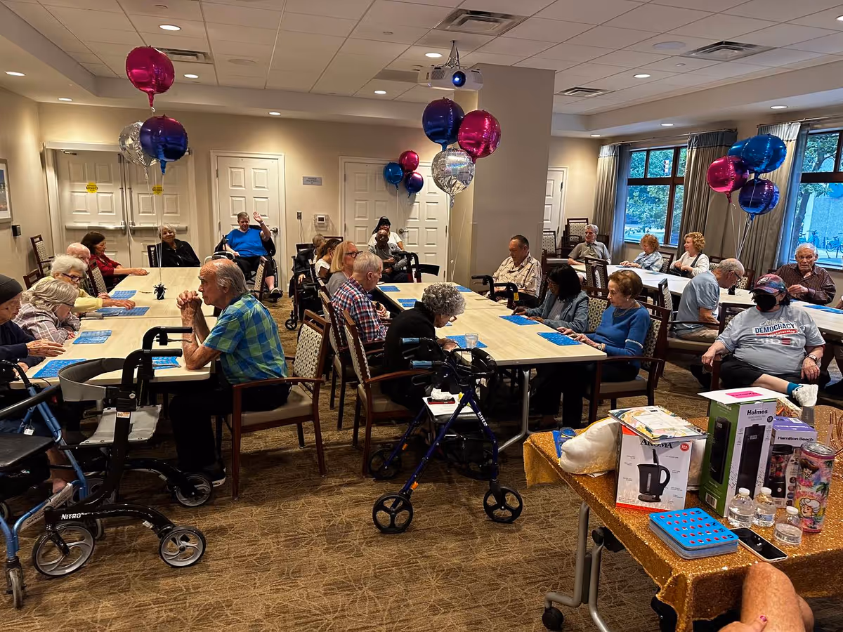 A group of elderly people seated around tables in a decorated room with balloons, some using walkers and wheelchairs, engaged in an activity or social event at Atria Center City.