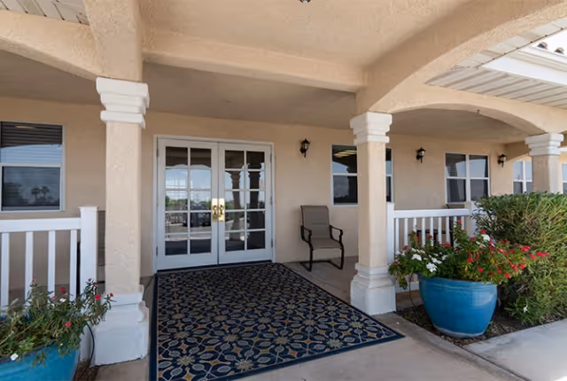 Covered entrance area of a senior living facility with double glass doors, a patterned outdoor rug, a single chair, white railing, and large blue planters with green and flowering plants.