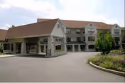 Exterior view of Dunwoody Village showing a multi-story building with a covered entrance driveway, surrounded by landscaped greenery and a paved driveway.