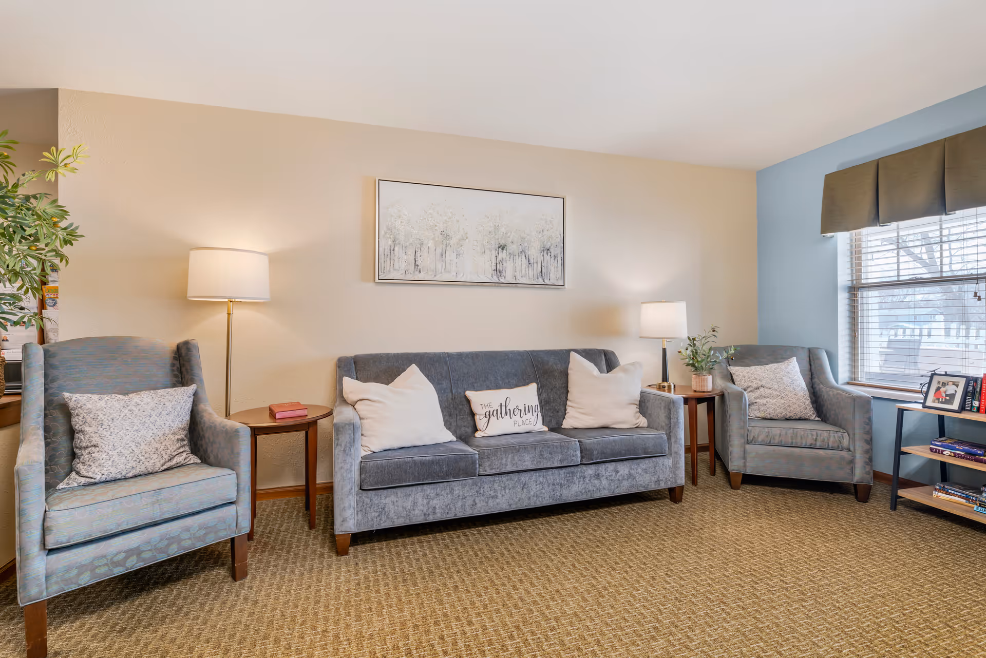 A cozy living room with a gray sofa and two matching armchairs, each with decorative pillows. There are two side tables with lamps on either side of the sofa, a framed artwork of trees on the wall above the sofa, a potted plant, and a window with blinds and a valance letting in natural light. The carpet is beige with a textured pattern.