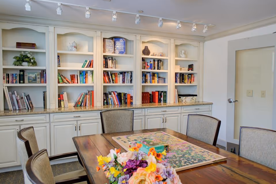A cozy room with a wooden table and six upholstered chairs. On the table, there is a partially completed jigsaw puzzle and a colorful bouquet of flowers. Behind the table, there is a built-in white bookshelf filled with books and decorative items. The room has a door with frosted glass on the right side and track lighting on the ceiling.