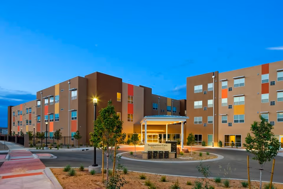 Exterior view of a modern multi-story senior living facility building at dusk with colorful panels on the facade, a circular driveway, landscaped plants, and a covered entrance with a sign that reads 'Supportive Care'.
