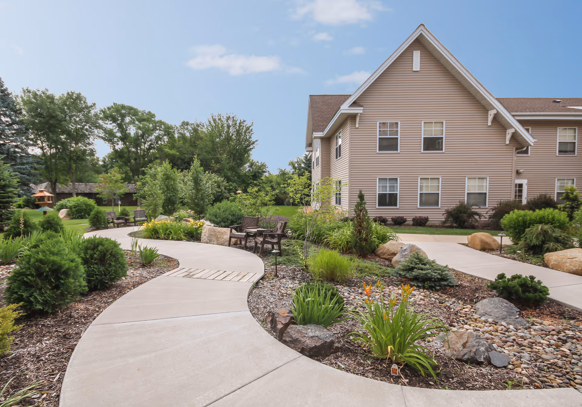 Curved concrete walkway surrounded by landscaped garden beds with rocks, shrubs, and flowers leading to benches and a beige two-story building under a blue sky with some clouds.