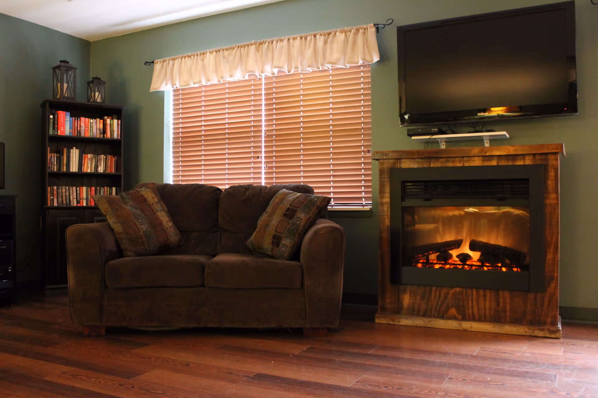 A cozy living room with a brown sofa adorned with patterned cushions, a wooden bookshelf filled with books, a window with closed blinds and a cream-colored valance, a wooden electric fireplace with a glowing fire, and a flat-screen TV mounted above the fireplace.