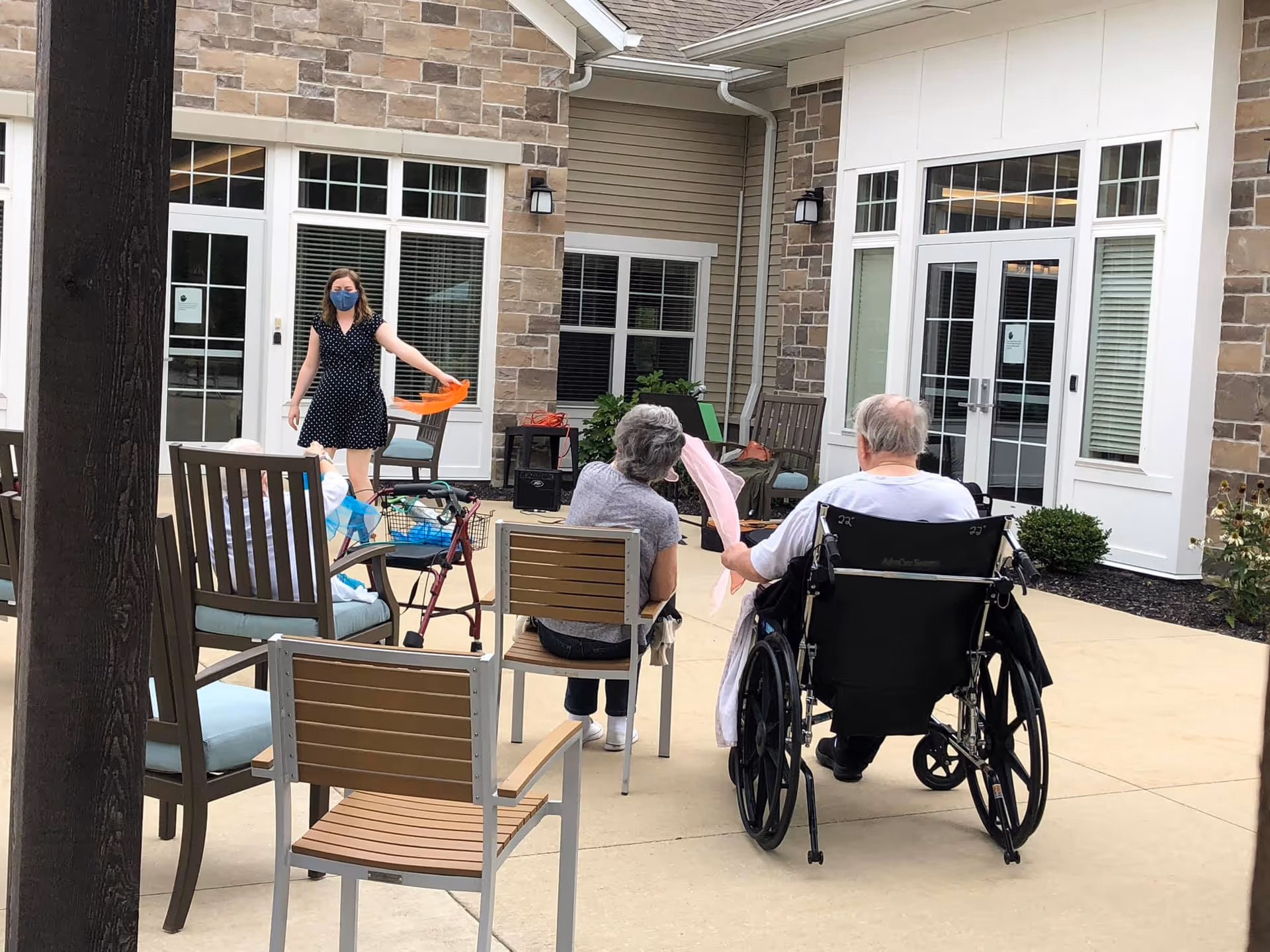 An outdoor courtyard area at a senior living facility with several elderly people seated on chairs and one person in a wheelchair. A woman wearing a mask and a black dress with white polka dots is standing and holding an orange object. The background shows a building with stone and siding exterior, white-framed windows, and glass doors.