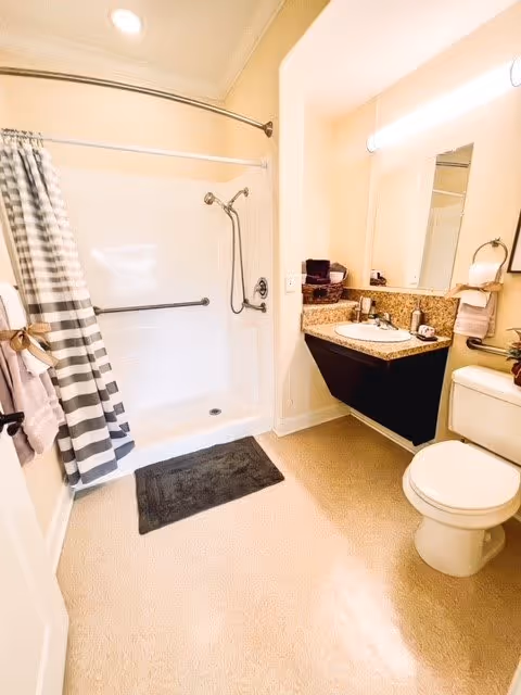 Bright bathroom with a white shower and a striped shower curtain, a black bath mat on the floor, a granite countertop with a sink, a mirror above the sink, and a toilet next to the sink. Towels are hanging on the wall and the door.