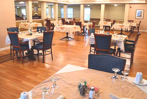 A spacious dining room with multiple tables covered in beige tablecloths, each set with glassware, napkins, and condiments. The room has wooden flooring and several windows allowing natural light to enter.