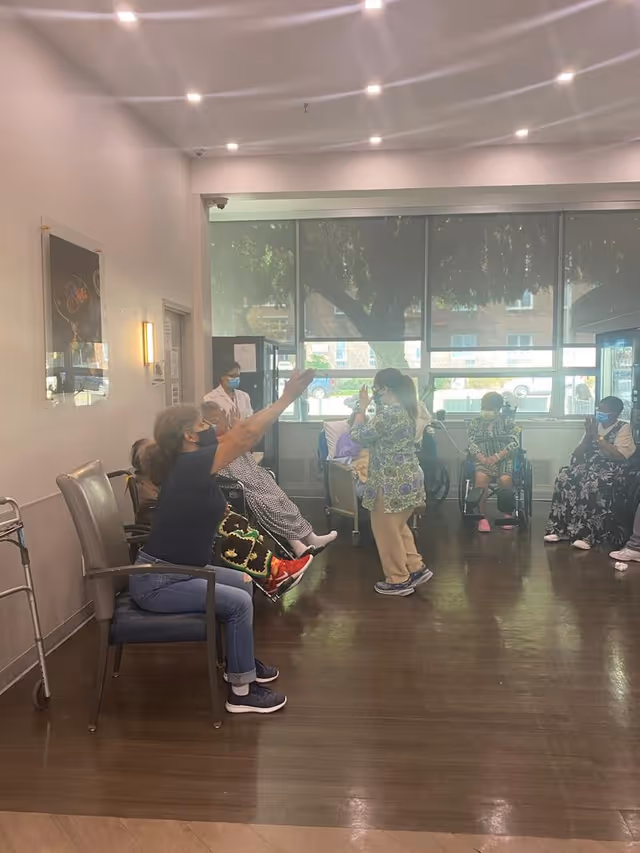 A group of elderly individuals and a caregiver wearing masks are engaged in an activity in a well-lit room with large windows. Some participants are seated in chairs or wheelchairs, while one person stands in the center. The room has wooden flooring and a walker is visible near the wall.