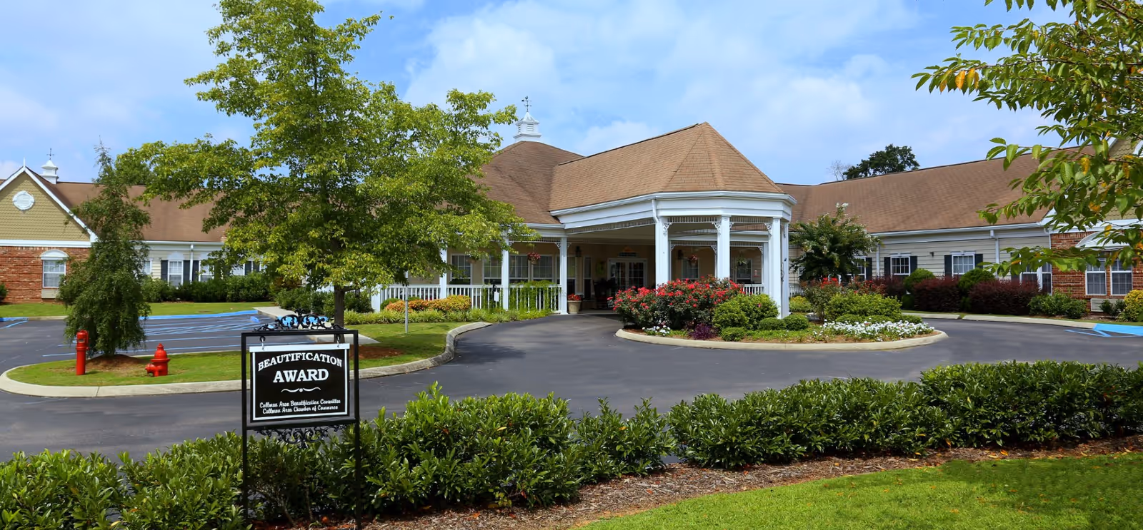 Front exterior view of The Neighborhood at Cullman senior living facility with a circular driveway, landscaped garden with flowers and shrubs, and a sign indicating a beautification award. The building has a covered entrance with white columns and a brown roof under a partly cloudy sky.