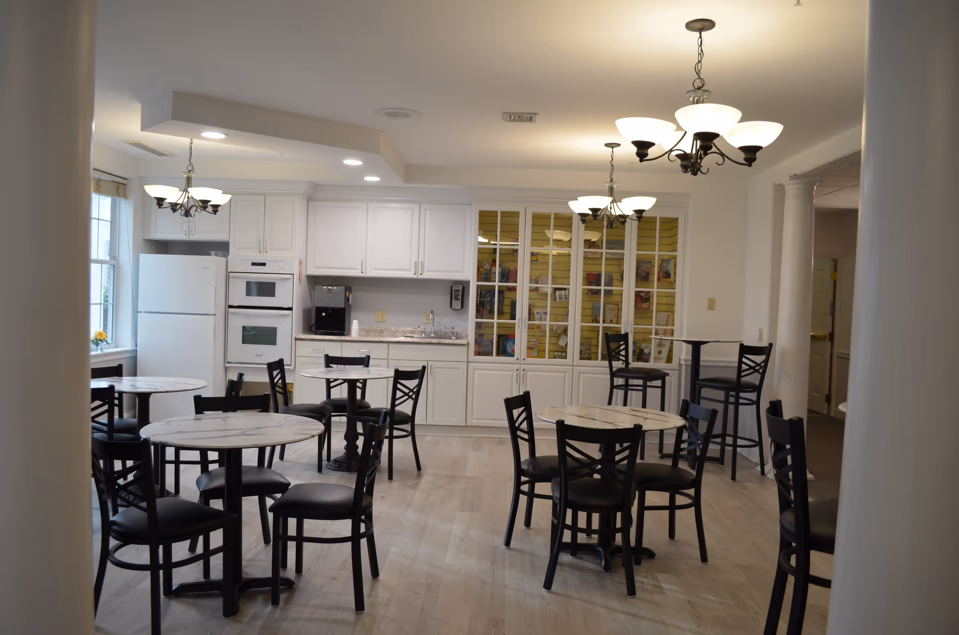 A bright dining area with several round tables and black chairs. The room features white cabinetry, a refrigerator, built-in oven, and a coffee machine. There are multiple chandeliers hanging from the ceiling, and a large window letting in natural light. The floor is light-colored wood, and there is a glass-front cabinet displaying various items.