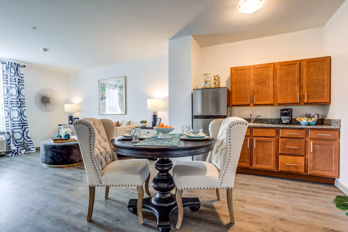 A bright and modern interior space featuring a small dining area with a round dark wooden table and four upholstered beige chairs. The table is set with plates, glasses, and a centerpiece bowl of oranges. Behind the dining area is a kitchenette with wooden cabinets, a granite countertop, a small refrigerator, and a coffee maker. To the left, there is a living area with a beige sofa, a round black ottoman, a side table with a lamp, and blue and white patterned curtains on the window.