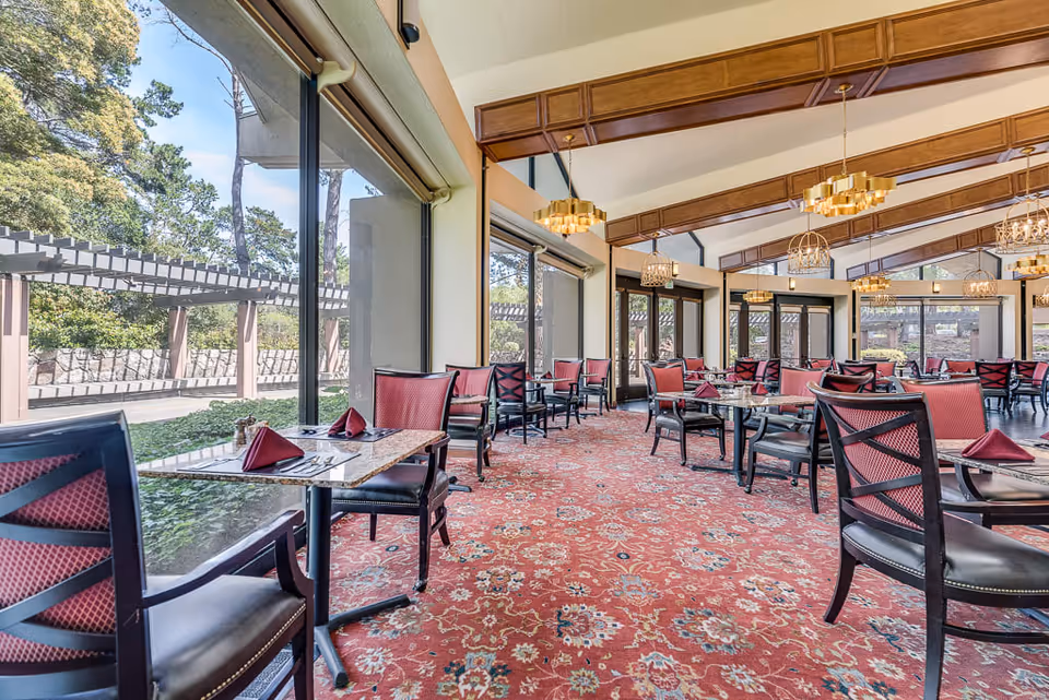 A spacious dining room with large windows offering a view of an outdoor garden area. The room features multiple tables set with folded red napkins, black chairs with red cushions, and a red patterned carpet. The ceiling has wooden beams and several modern chandeliers providing warm lighting.