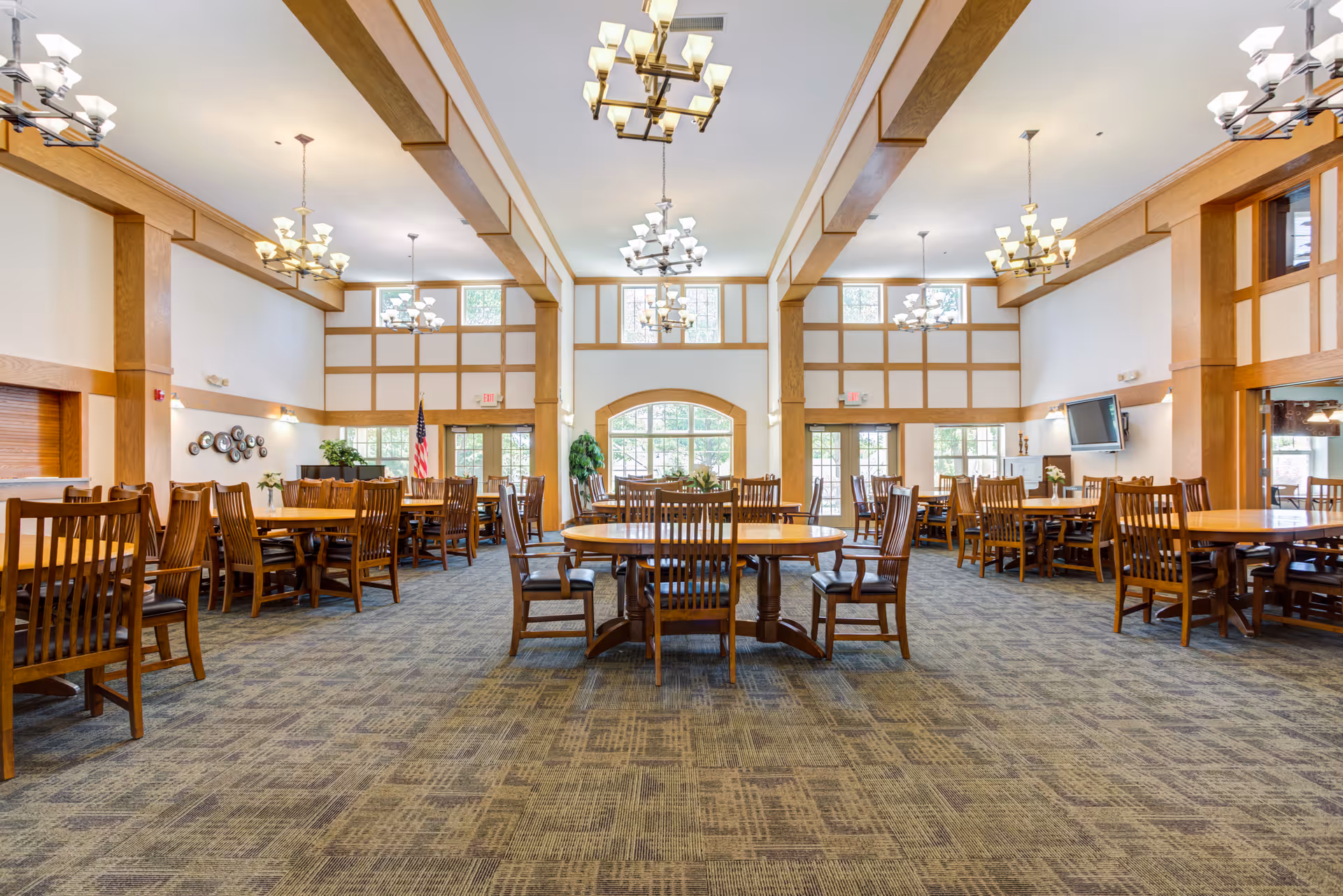 Spacious dining room with multiple wooden tables and chairs arranged neatly. The room features high ceilings with wooden beams, large windows allowing natural light, and several chandeliers hanging from the ceiling. There is an American flag near the back wall and a television mounted on the right wall.